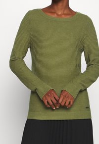 Sweter z dzianiny w kolorze oliwkowej zieleni o prążkowanej fakturze, z okrągłym dekoltem i długimi rękawami. Posiada małą metkę marki przy dolnym brzegu.