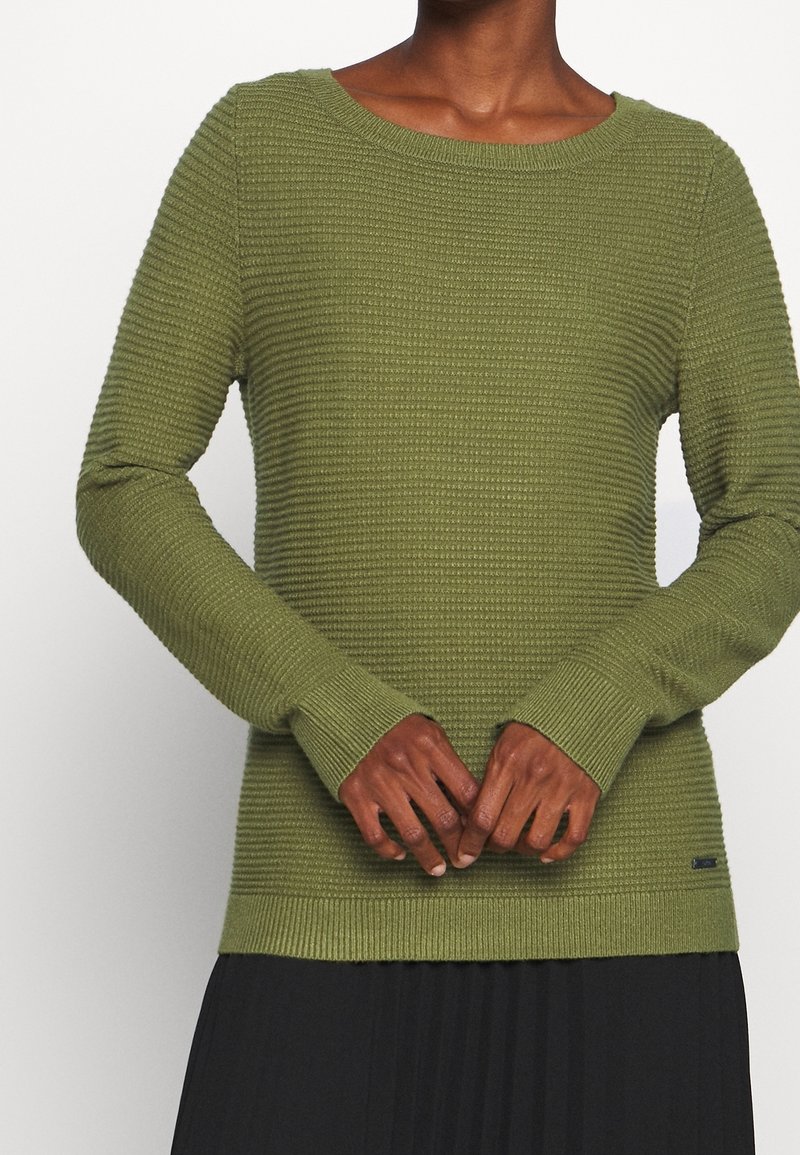 Sweter z dzianiny w kolorze oliwkowej zieleni o prążkowanej fakturze, z okrągłym dekoltem i długimi rękawami. Posiada małą metkę marki przy dolnym brzegu.