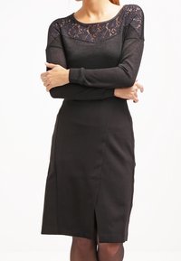 Black dress featuring long sleeves and a lace bodice. The fitted design extends to knee length with a subtle front slit. Smooth texture overall.