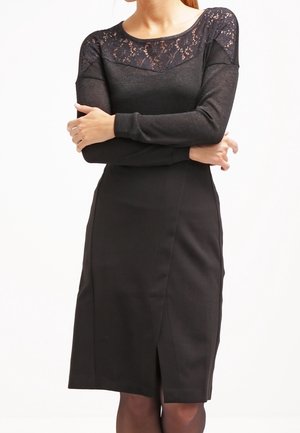 Robe noire avec des manches longues et un corps en dentelle. La coupe ajustée s'étend jusqu'au genou avec une fente discrète à l'avant. Texture lisse dans l'ensemble.