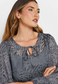 Blusa grigia trasparente con stampa animale testurizzata, scollatura a chiave con chiusura a fiocco e maniche ampie. Leggero luccichio aggiunge profondità.