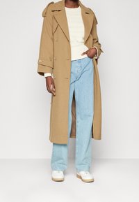 Cappotto cammello con bottoni doppi, ampie rever e spalline, abbinato a jeans azzurri chiari e sneakers bianche con zeppa.
