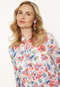 Una donna con capelli biondi e ondulati indossa una blusa floreale con motivi rossi, blu e bianchi e grandi orecchini, sorridendo dolcemente su uno sfondo chiaro.