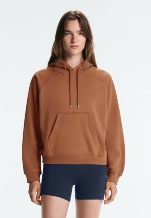 Jeune femme aux longs cheveux bruns portant un sweat à capuche marron et un short moulant bleu foncé, debout devant un fond uni.