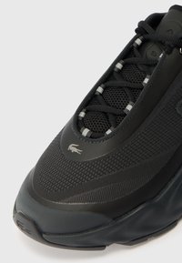 Černá atletická bota s vrškem z prodyšné síťoviny, texturovanými panely a šněrovacím designem. Obsahuje gumovou podrážku a malé logo blízko špičky.