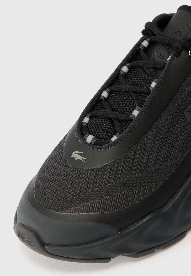 Schwarzer Sportschuh mit Mesh-Obermaterial, strukturierten Einsätzen und Schnürdesign. Verfügt über eine Gummisohle und ein kleines Logo nahe der Zehenpartie.