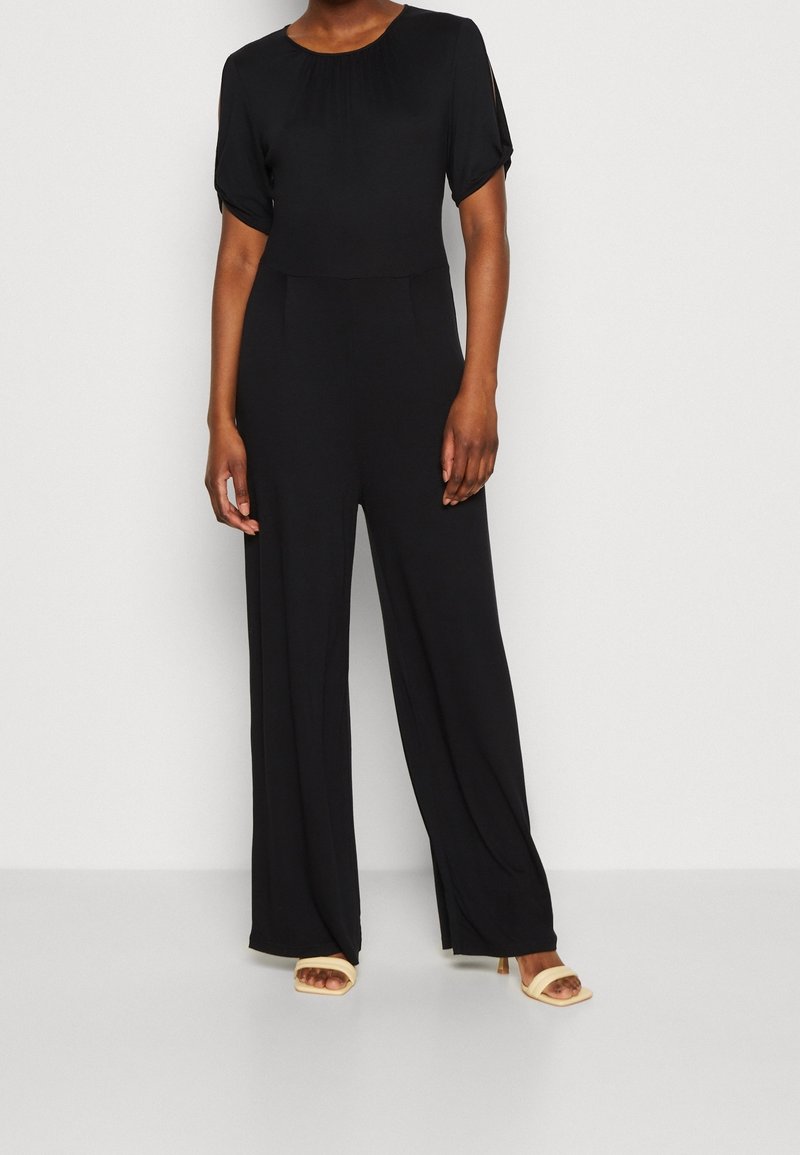 Černý overall s krátkými, nabíranými rukávy, kulatým výstřihem a širokými kalhotami. K tomu jsou obuty béžové sandály na podpatku. Měkká tkanina, splývavý design.