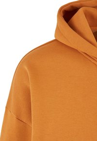 Haut à capuche orange en tissu doux et texturé, avec une grande capuche et une coupe décontractée aux épaules tombantes. Aucun motif ni matériel visible.