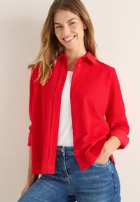 Röd skjorta med krage, uppvikta ärmar, gjord av texturerat tyg, i kombination med en vit topp och jeans.