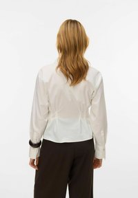 Weiße Bluse mit langen Ärmeln, Rückenfalten und geknöpften Manschetten, kombiniert mit dunkler Hose. Glatter Stoff, maßgeschneiderte Passform, minimalistisches Design.