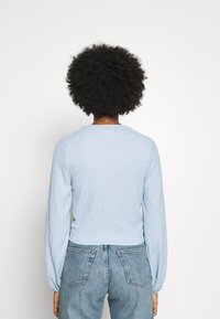 Person med lockigt hår som bär en ljusblå långärmad topp och högmidjade blå jeans, står vänd mot en vit bakgrund.