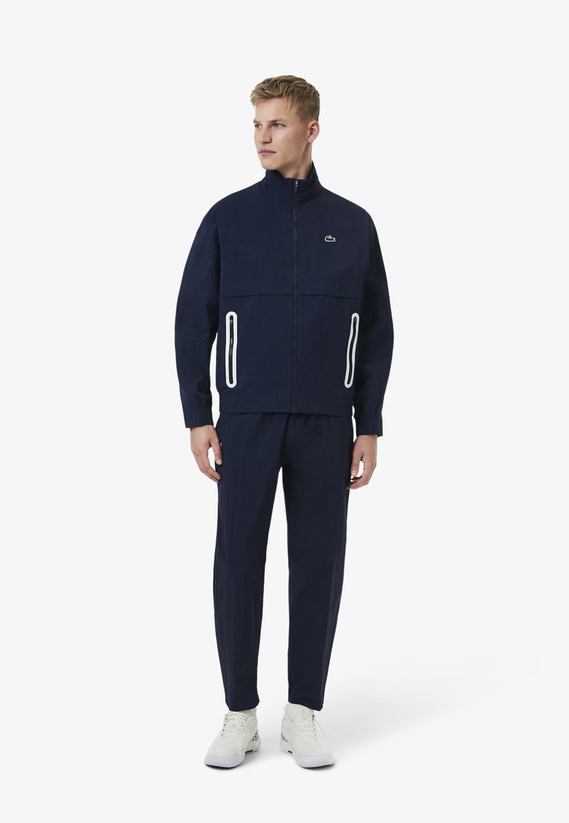 Homme debout portant un survêtement bleu marine avec une veste à fermeture éclair, des poches zippées soulignées en blanc et des baskets blanches, regardant sur le côté.