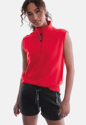 Junge Frau trägt ein ärmelloses rotes Oberteil mit Reißverschluss und schwarze Shorts mit Kordelzug, steht vor einem schlichten weißen Hintergrund.
