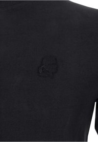Camiseta de algodón negra con un logo bordado texturizado del mismo color en el lado izquierdo del pecho. La tela tiene un acabado suave.