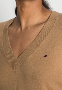 V-pääntiellä varustettu hiekanvärinen neulepaita, jossa on ribbineulotut reunat ja pieni kirjailtu logo rinnassa punaisen, valkoisen ja sinisen sävyissä. Pehmeä tekstuuri.