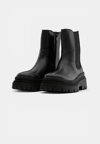 Paire de bottines en cuir noir avec semelles crantées épaisses et fermetures éclair latérales sur fond blanc.