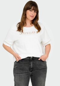 Witte katoenen t-shirt met "Amour" in een gestreept patroon gedrukt, met korte mouwen met oogjesdetails. Gecombineerd met donkergrijze jeans.