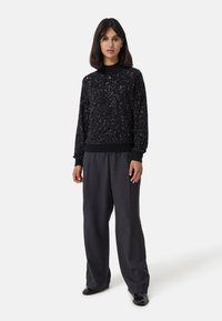 Maglione nero con paillettes, collo rotondo, vestibilità comoda e maniche lunghe, abbinato a pantaloni grigi scuri larghi e scarpe nere.