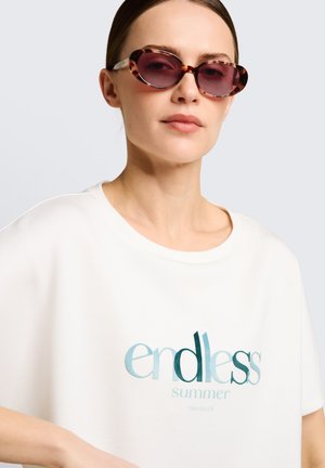Femme portant des lunettes de soleil ovales en écaille de tortue et une chemise blanche portant l'inscription « endless summer » sur un fond gris clair.