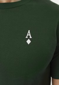 Σκούρο πράσινο μπλουζάκι με λευκό γράμμα "A" πάνω από ένα διαμάντι τυπωμένο στην αριστερή πλευρά του στήθους.