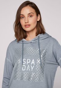 Felpa con cappuccio azzurro chiaro caratterizzata da una stampa metallica argento "SPA DAY" circondata da un motivo testurizzato. Tessuto morbido con cordini.