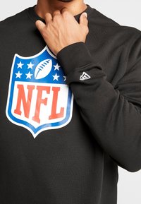 Felpa nera con logo NFL che presenta uno scudo blu, scritte rosse e una grafica di un pallone da football. Polsini a coste e logo discreto sulla manica.