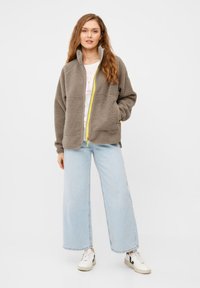 Graue Fleecejacke mit hohem Kragen und gelbem Reißverschluss, über einem weißen T-Shirt getragen, kombiniert mit hellblauen, weiten Jeans und weißen Sneakers.