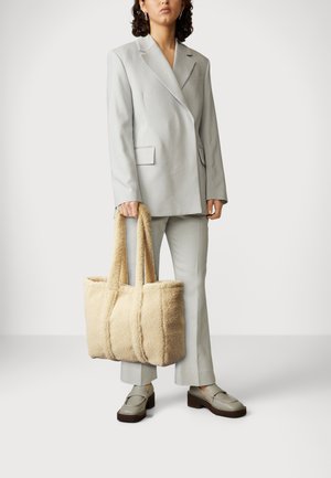 Beige shearling-tote taske med blød tekstur, to robuste håndtag og et struktureret design. Bæres med en lysegrå dragt.