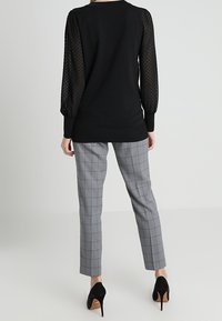 Musta neule, jossa on teksturoidut, pilkulliset läpikuultavat hihat, yhdistetty harmaisiin ruudullisiin housuihin ja mustiin korkokenkiin.