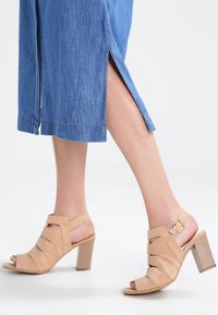 Nainen, joka käyttää beigen värisiä remmikäynnöksiä korkokenkiä yhdistettynä siniseen farkkuhameeseen, jossa on sivureikä.