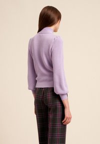 Fialový žebrovaný svetr s vysokým límcem a nařasenými rukávy, doplněný kostkovanými kalhotami s černými, zelenými a fialovými pruhy.