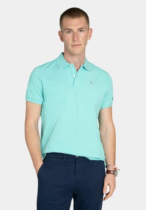 Giovane uomo con i capelli corti che indossa una polo color turchese chiaro e pantaloni blu scuro, in piedi con una mano in tasca.