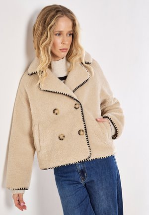 Veste en shearling beige à double boutonnage avec des coutures noires, de grands boutons, des poches latérales et un large col, associée à un jean bleu.