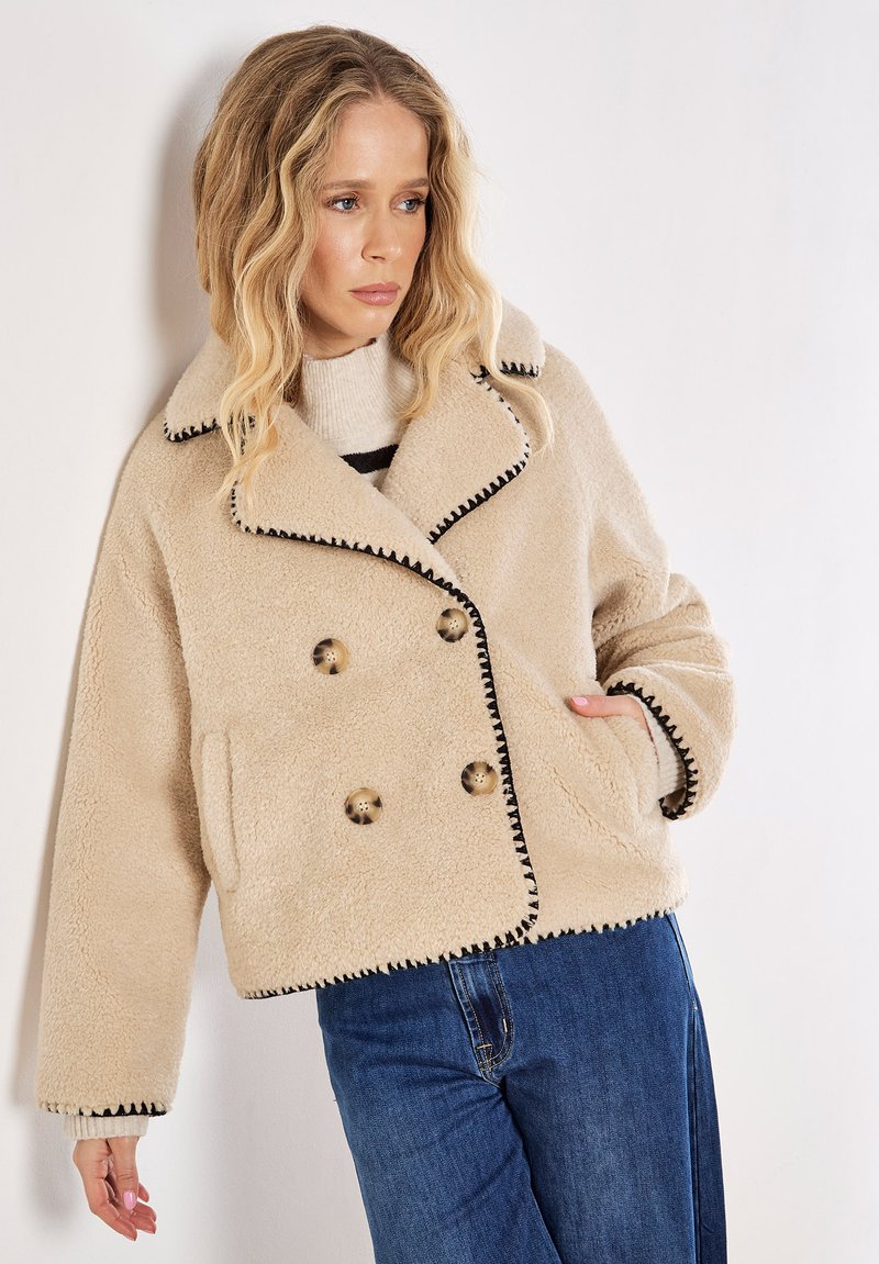 Beige dubbelzijdig shearling jasje met zwarte stiksels, grote knopen, zijzakken en een brede kraag, gecombineerd met een blauwe spijkerbroek.