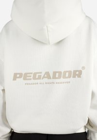 Biely mikina s kapucňou s veľkým béžovým logom "PEGADOR" potlačeným na zadnej strane. Textúrovaná tkanina, obsahuje kapucňu a pruhovaný lem.