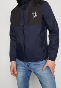 Giacca a vento blu navy con pannello nero, chiusura con zip, grafica del logo, polsini elasticizzati e cappuccio. Materiale liscio e leggero.