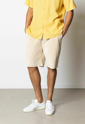 Lys gul kortærmet skjorte med et struktureret mønster, kombineret med beige kordfløjls shorts. Hvide sneakers fuldender outfittet.