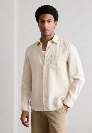 Ung mand med kort krøllet hår iført en beige skjorte med knapper over en hvid undertrøje og beige bukser, stående med den ene hånd i lommen.