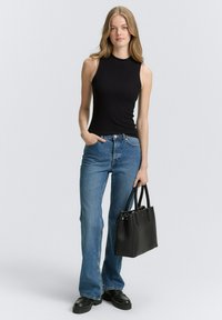 Schwarzes ärmelloses Rippoberteil, kombiniert mit blauen Schlagjeans. Das Model hält eine schwarze Lederhandtasche mit Henkeln. Die Schuhe sind klobige schwarze Stiefel.