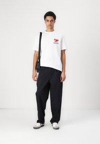 Hvid bomulds t-shirt med rød grafisk design og tekst, parret med løse sorte bukser og grå sneakers. Sort crossover-taske som tilbehør.