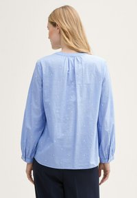 Blouse bleu clair avec des manches longues à volants et de subtils pois brodés blancs. Tissu en coton lisse avec une coupe décontractée et un col rond.