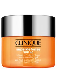 Oransje glasskrukke med lokk i sølvmetall, merket "Clinique Superdefense SPF 40." Merkbare tekster inkluderer bruksområde for tretthet og aldringstegn.