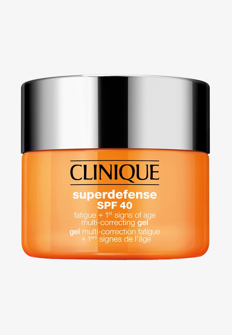 Oransje glasskrukke med lokk i sølvmetall, merket "Clinique Superdefense SPF 40." Merkbare tekster inkluderer bruksområde for tretthet og aldringstegn.