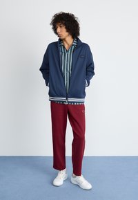 Navy zip-up jacket with white stripes, layered over a green-striped shirt, paired with burgundy pants and white sneakers.