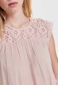 Blusa rosa claro con un yugo de encaje, escote redondeado y cuerpo plisado. La tela tiene un acabado suave y texturizado.