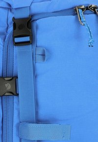 Pochette en tissu bleu avec une boucle en plastique noir, des sangles bleues et un détail en cordon. La texture semble lisse avec une construction robuste.