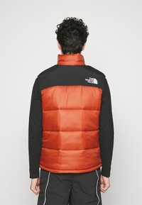 Červená a černá zateplená puffer vesta s vysokým límcem, která má horizontální prošívání, logo na zadní straně a hladkou texturu.