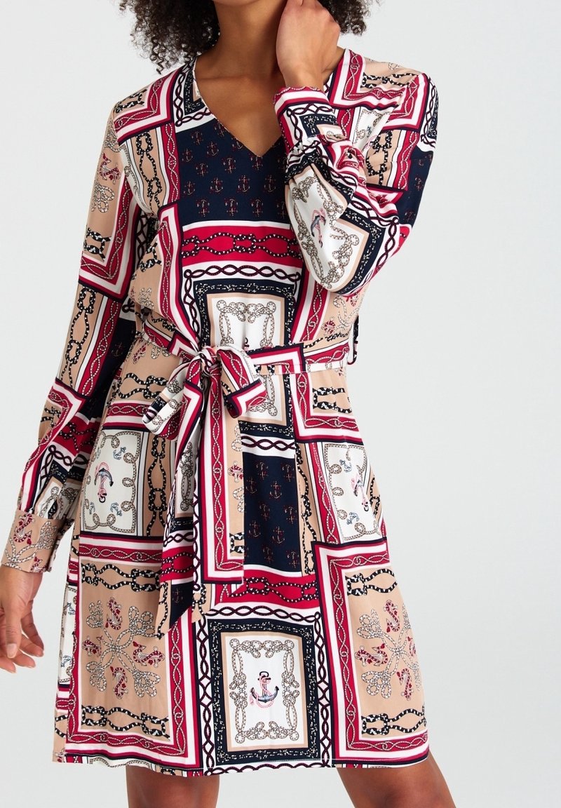 Femme portant une robe ceinturée à manches longues avec un imprimé géométrique et chaîne rouge, marine et beige, debout devant un fond clair uni.