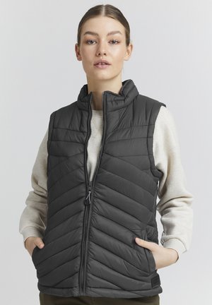 Gilet matelassé gris avec un col montant, présentant un piquage diagonal, une fermeture éclair à l'avant et des poches latérales. Porté par-dessus un pull léger.
