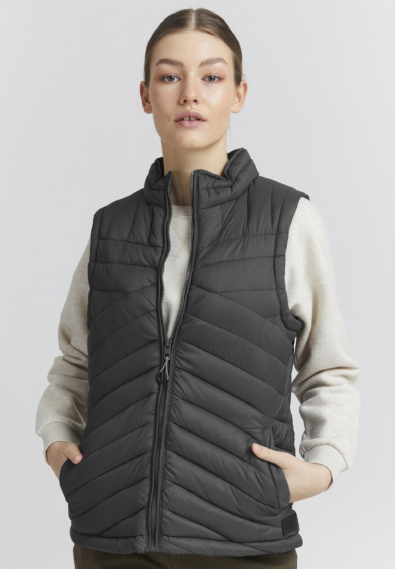 Gilet imbottito grigio con colletto alto, caratterizzato da un'acopertura diagonale, una cerniera frontale e tasche laterali. Indossato sopra un maglione leggero.